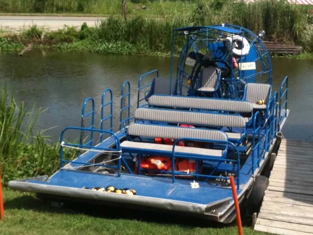 A blue airboat with four long bench rows and a caged propeller, tied up at a wooden dock on a bayou canal.